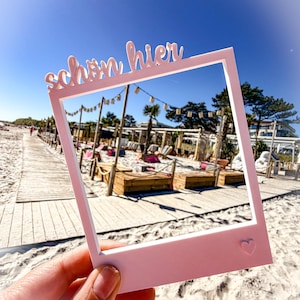May include: A pink photo frame with the German text "sch&ouml;n hier" and a heart symbol. The frame is held up against a beach scene with a wooden boardwalk, sand, and a clear blue sky.