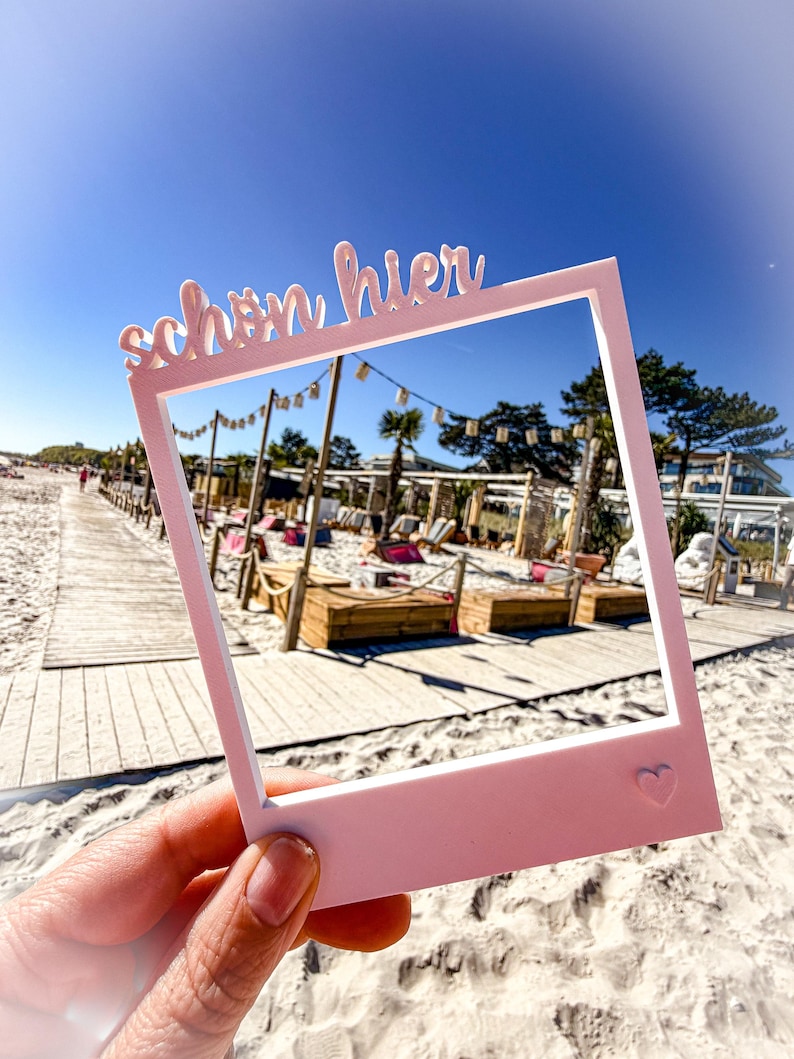 May include: A pink photo frame with the German text "sch&ouml;n hier" and a heart symbol. The frame is held up against a beach scene with a wooden boardwalk, sand, and a clear blue sky.