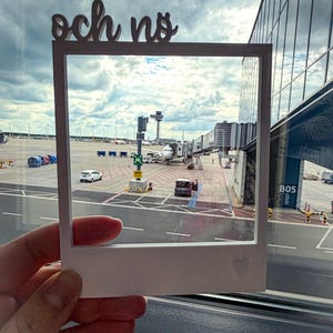 May include: A white, Polaroid-style frame with the words "och no" cut out at the top. The frame is held up to a window, showing an airport scene with airplanes, terminals, and a cloudy sky. A small heart is etched on the frame.