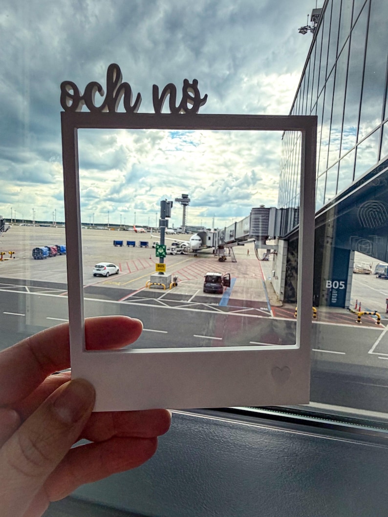 May include: A white, Polaroid-style frame with the words "och no" cut out at the top. The frame is held up to a window, showing an airport scene with airplanes, terminals, and a cloudy sky. A small heart is etched on the frame.