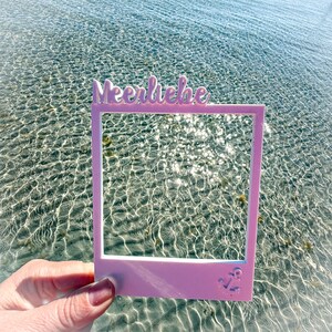 May include: A light pink photo frame with the word "Meerliebe" in cursive script at the top. An anchor is at the bottom right corner of the frame. The frame is held in front of a body of water.