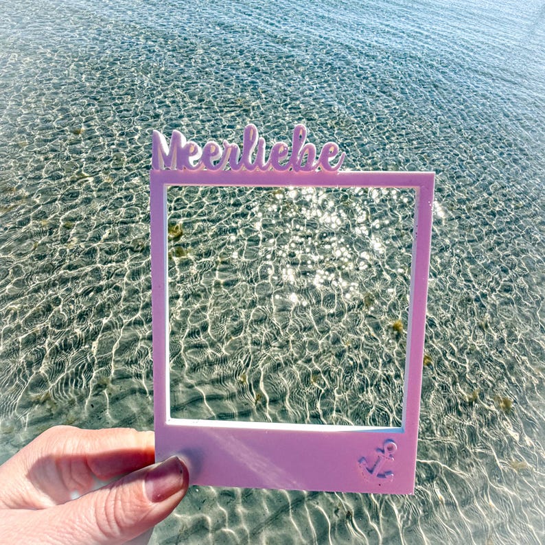 May include: A light pink photo frame with the word "Meerliebe" in cursive script at the top. An anchor is at the bottom right corner of the frame. The frame is held in front of a body of water.
