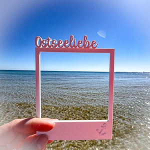 May include: A pink rectangular frame with the word "Ostseeliebe" in cursive script across the top. An anchor is etched in the bottom right corner. The frame is held up against a backdrop of the ocean and a clear blue sky.
