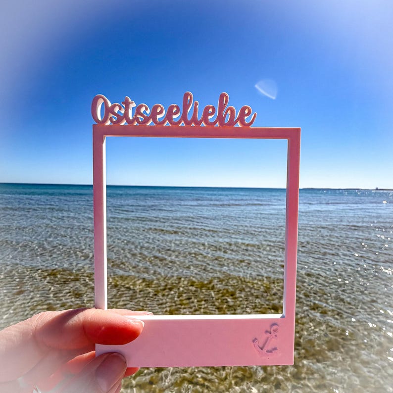 May include: A pink rectangular frame with the word "Ostseeliebe" in cursive script across the top. An anchor is etched in the bottom right corner. The frame is held up against a backdrop of the ocean and a clear blue sky.