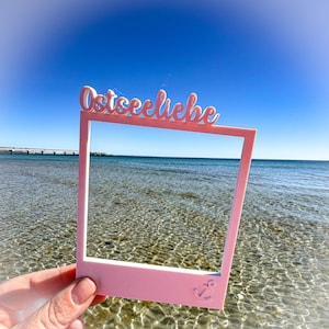 May include: A pink photo frame with the word "Ostseeliebe" in cursive script across the top. The frame has an anchor design in the lower right corner. The frame is held up against a backdrop of the ocean and a clear blue sky.