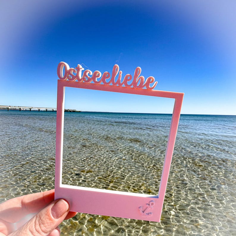 May include: A pink photo frame with the word "Ostseeliebe" in cursive script across the top. The frame has an anchor design in the lower right corner. The frame is held up against a backdrop of the ocean and a clear blue sky.