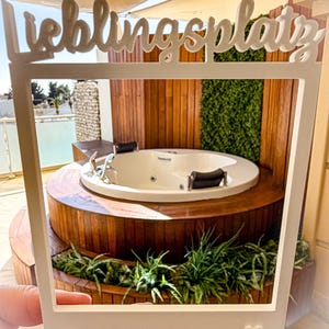 May include: A white photo frame with the word "Lieblingsplatz" in script, held in front of a hot tub. The hot tub is white, surrounded by wooden decking and greenery. A small heart is on the frame.