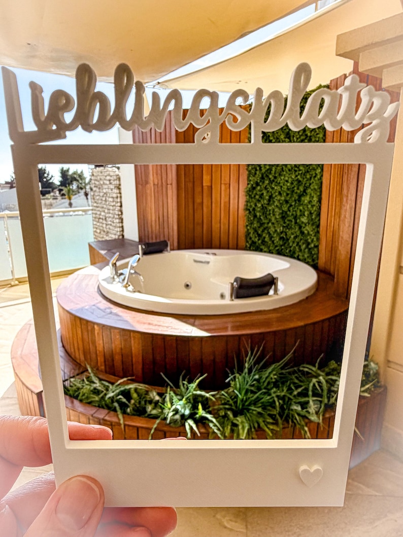 May include: A white photo frame with the word "Lieblingsplatz" in script, held in front of a hot tub. The hot tub is white, surrounded by wooden decking and greenery. A small heart is on the frame.