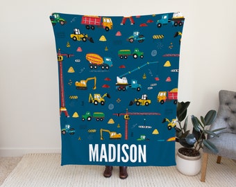 Op de afbeelding: Een blauwe fleece deken met een patroon van bouwvoertuigen en de naam "MADISON" in wit gedrukt.
