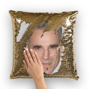 May include: A gold sequin pillow with a man's face peeking out from behind the sequins. The face is visible in the center of the pillow. The sequins are gold and white.