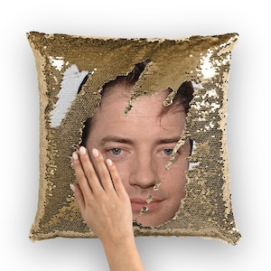 May include: A square pillow with gold sequins, revealing a portrait of a person. The sequins are partially flipped to show a white section. A hand is touching the portrait. The pillow is on a white background.