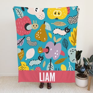 May include: A blue and pink personalized throw blanket with a colorful pattern of apples, pears, leaves, and caterpillars. The blanket has a red border with the name "LIAM" printed in white.