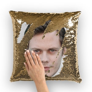 May include: A gold sequin pillow with a man's face peeking out from behind the sequins. The face is visible when the sequins are moved aside.