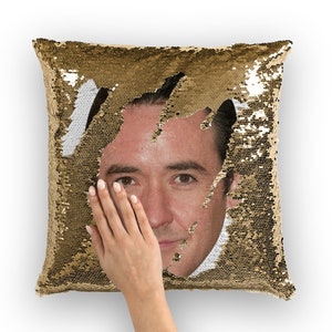 May include: A gold sequin pillow with a man's face peeking out from behind the sequins. The face is visible through a tear in the sequins.