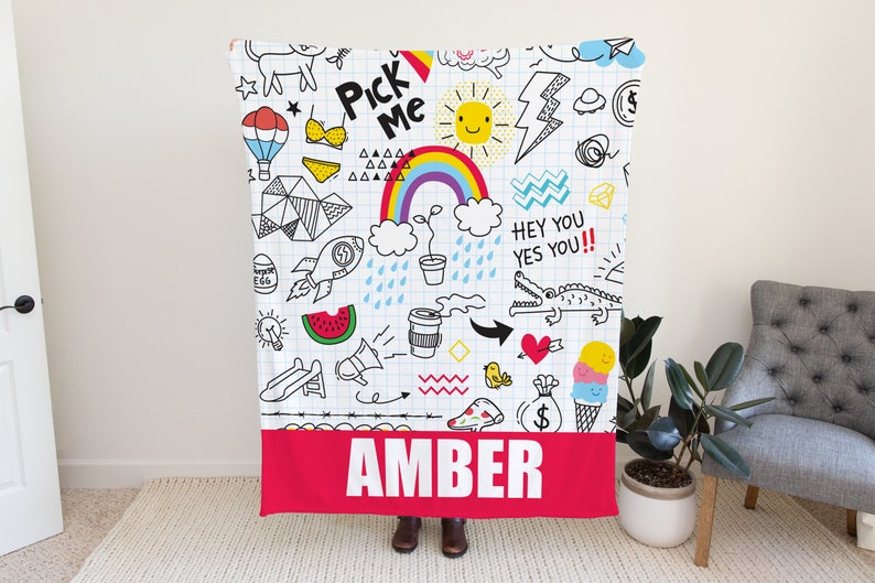 May include: A white fleece blanket with a colorful doodle design. The doodles include a rainbow, a sun, a watermelon, a crocodile, and a hot air balloon. The blanket has a red border with the name "AMBER" printed in white.