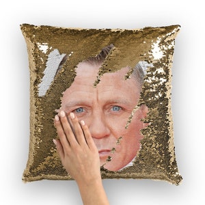 Peut inclure: Un coussin carré décoratif avec des paillettes dorées. Une main recouvre partiellement un portrait d'un homme aux yeux bleus. Les paillettes peuvent être retournées pour révéler une surface blanche.