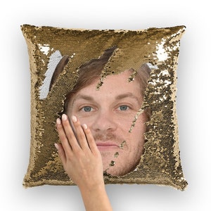 Op de afbeelding: Een kussen van gouden pailletten met een mannengezicht dat uit het midden kijkt. Het gezicht is zichtbaar door een gat in de pailletten. Het kussen is zacht en pluche.