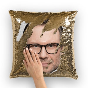 Op de afbeelding: Een kussen van gouden sequins met een mannengezicht dat tevoorschijn komt achter de sequins. De man draagt een bril en heeft een baard.