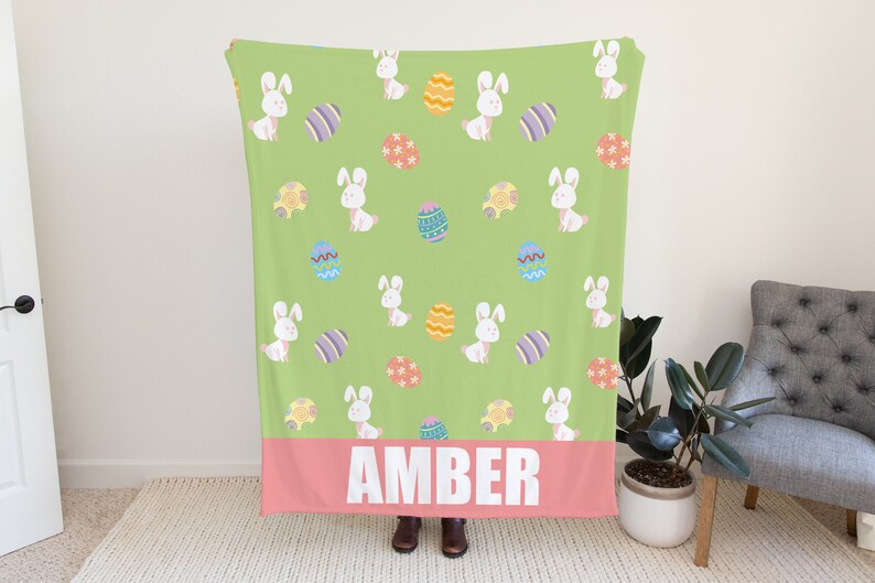 Puede incluir: Una manta de forro polar verde con un patr&oacute;n repetido de conejos blancos y huevos de Pascua coloridos. La manta tiene un borde rosa con el nombre "AMBER" impreso en blanco.