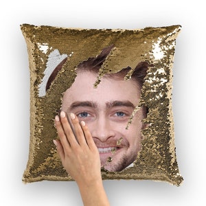 Peut inclure: Un oreiller en sequins dorés avec un visage d'homme qui apparaît au centre. Le visage est souriant et a une barbe.