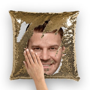 Puede incluir: Una almohada de lentejuelas doradas con la cara de un hombre asomando entre las lentejuelas. La cara sonríe y las lentejuelas son movidas por una mano.