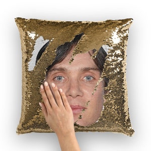 May include: Gold sequin pillow with a portrait revealed in the center. The pillow's sequins are gold, and the revealed image shows a person with blue eyes. A hand is touching the image.