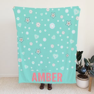 May include: A turquoise blue fleece blanket with a white and pink floral pattern and the name "AMBER" printed in pink on the bottom.