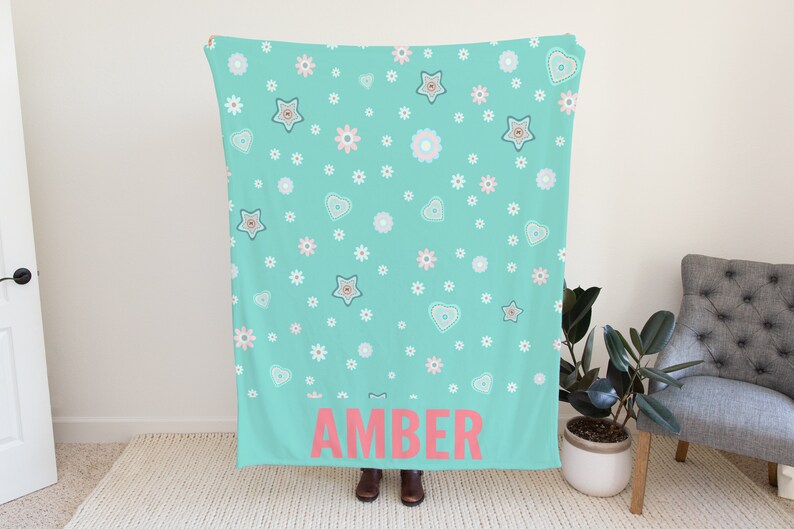 May include: A turquoise blue fleece blanket with a white and pink floral pattern and the name "AMBER" printed in pink on the bottom.