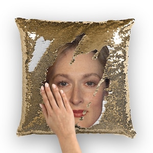 May include: A gold sequin pillow with a woman's face peeking through a hole in the sequins. The woman is looking down with a sad expression.