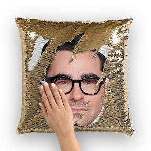 May include: A gold sequin pillow with a man's face peeking out from behind the sequins. The man is wearing glasses and has a serious expression.