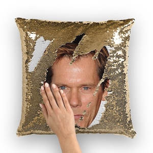 May include: A gold sequin pillow with a man's face peeking out from a hole in the sequins. The man is looking at the camera with a serious expression.