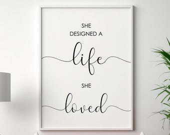 Puede incluir: Una impresión enmarcada en blanco con texto negro que dice "She designed a life she loved" colgada en una pared blanca sobre un sofá rojo con almohadas blancas. Una planta en maceta está en el lado derecho de la imagen.