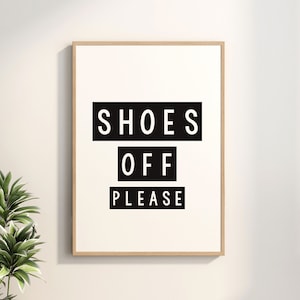 May include: A framed print with the text "SHOES OFF PLEASE" in a bold, black font on a white background. The frame is a light wood color. A potted plant is visible in the lower left corner.