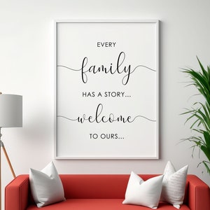 May include: A white framed print with black text that reads "Every family has a story... welcome to ours..." The print is hanging on a white wall above a red sofa with white cushions.