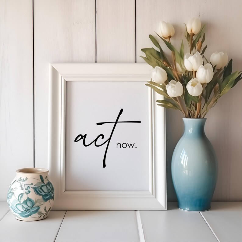 May include: White framed print with black calligraphy that reads "act now." The print is sitting on a white surface next to a white ceramic vase with white flowers and a small blue and white ceramic vase.