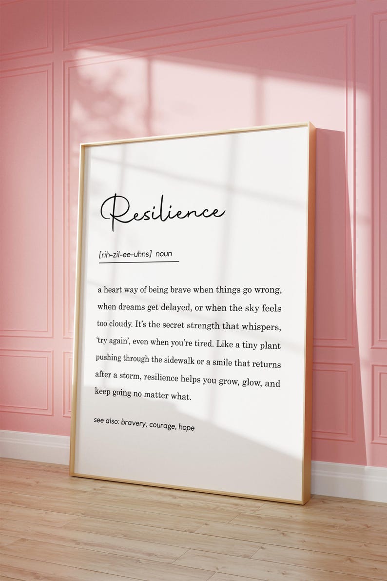 May include: A framed print with the definition of "Resilience" in elegant script. The definition is written in black text on a white background, with a gold frame. The print is leaning against a pink wall.