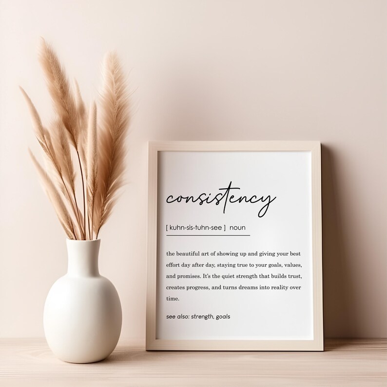 May include: A framed print with a white background and black text. The text reads "consistency" and defines the word as "the beautiful art of showing up and giving your best effort day after day, staying true to your goals, values, and promises. It's the quiet strength that builds trust, creates progress, and turns dreams into reality over time." The text also includes "see also: strength, goals". The print is sitting on a light brown wooden surface with a vase of dried pampas grass.