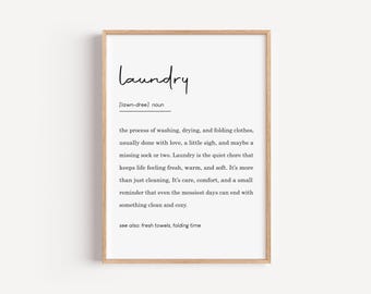 May include: A framed print with the word "Laundry" in a handwritten style font. The definition of laundry is written below, describing the process of washing, drying, and folding clothes. The frame is a light brown color.