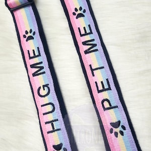 May include: Two black pet carrier straps with rainbow stripes. One strap says "HUG ME" and the other says "PET ME" in black text. Each strap has a black paw print. The straps have black plastic buckles.