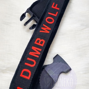 May include: A black fabric collar featuring the text DUMB WOLF embroidered in bright red capital letters, flanked by two red paw print icons. The collar includes a black plastic side-release buckle mechanism for fastening and adjustment.