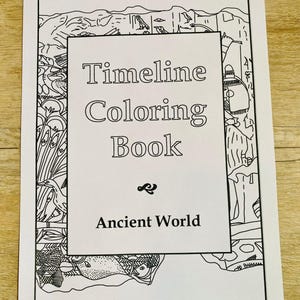 Op de afbeelding: Een zwart-wit kleurboek getiteld "Timeline Coloring Book Ancient World". De omslag heeft een decoratieve rand met illustraties in oude Egyptische stijl. Het boek is gedrukt op wit papier en ligt op een lichtbruin houten oppervlak.