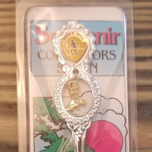 May include: A silver souvenir spoon with a gold-colored Boot Hill, Kansas emblem. The spoon is packaged in a clear plastic sleeve with a colorful card featuring a red sun and a green tree.