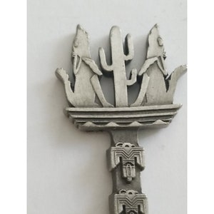 May include: A silver key with a design of three coyotes howling at a cactus. The key has a decorative stem with a repeating pattern of stylized birds.