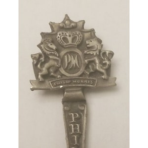 May include: Silver spoon with a Philip Morris logo. The logo features a crown, two lions, and the letters PM. The spoon handle has the word "PHILIP" engraved on it.