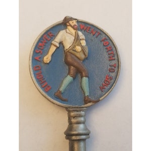 May include: A silver key with a blue and white round metal charm. The charm features a cartoon illustration of a man walking with a bag over his shoulder. The text on the charm reads "Behold a Sower Went Forth to Sow".