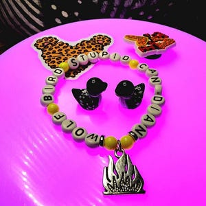 May include: A circular beaded bracelet featuring white letter beads spelling STUPID BIRD CANADIAN WOLF, interspersed with yellow round beads. A silver-toned metal flame charm hangs from the bottom. Two small black duck figurines are inside the circle.