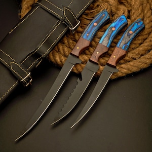 May include: Three knives with matte black blades and handles featuring a combination of dark brown wood and blue-dyed wood grain. Each handle is secured with three brass rivets. The knives are positioned next to a black leather sheath.