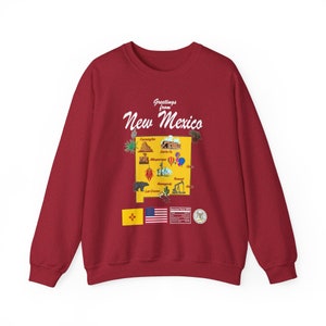 Pode incluir: Camisola vermelha com um gráfico branco do estado do Novo México. O gráfico inclui o texto "Greetings from New Mexico" e um mapa do estado com vários marcos e cidades rotulados. O gráfico também inclui a bandeira do estado do Novo México e a bandeira americana.