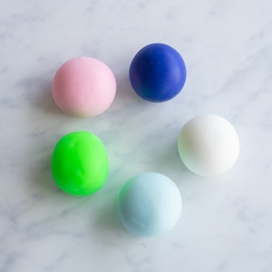 May include: Six colourful bouncy balls in different colours: pink, blue, white, green, and light blue. The balls are arranged in a circle on a white marble surface.