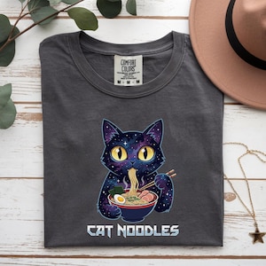 May include: Dark grey t-shirt featuring a cartoon cat with a galaxy design eating ramen noodles from a bowl. The text "CAT NOODLES" is printed below the cat in a stylized font. The shirt has a Comfort Colors tag.